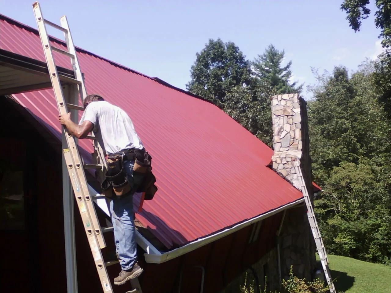 Roofer on a ladder beside a red metal roof and stone chimney — Soffit & Fascia Repair by Roofing Brothers in Granite Quarry